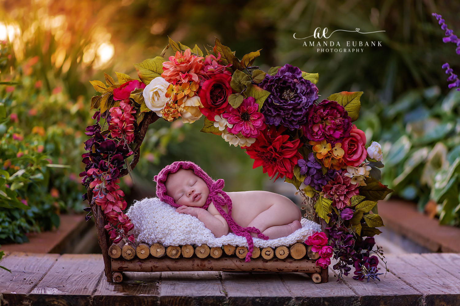 Destin Newborn Photographer | Floral Arch Newborn Session Destin newborn photographer captures a sleeping baby girl in a pink bonnet under a vibrant floral arch on a wooden bed.